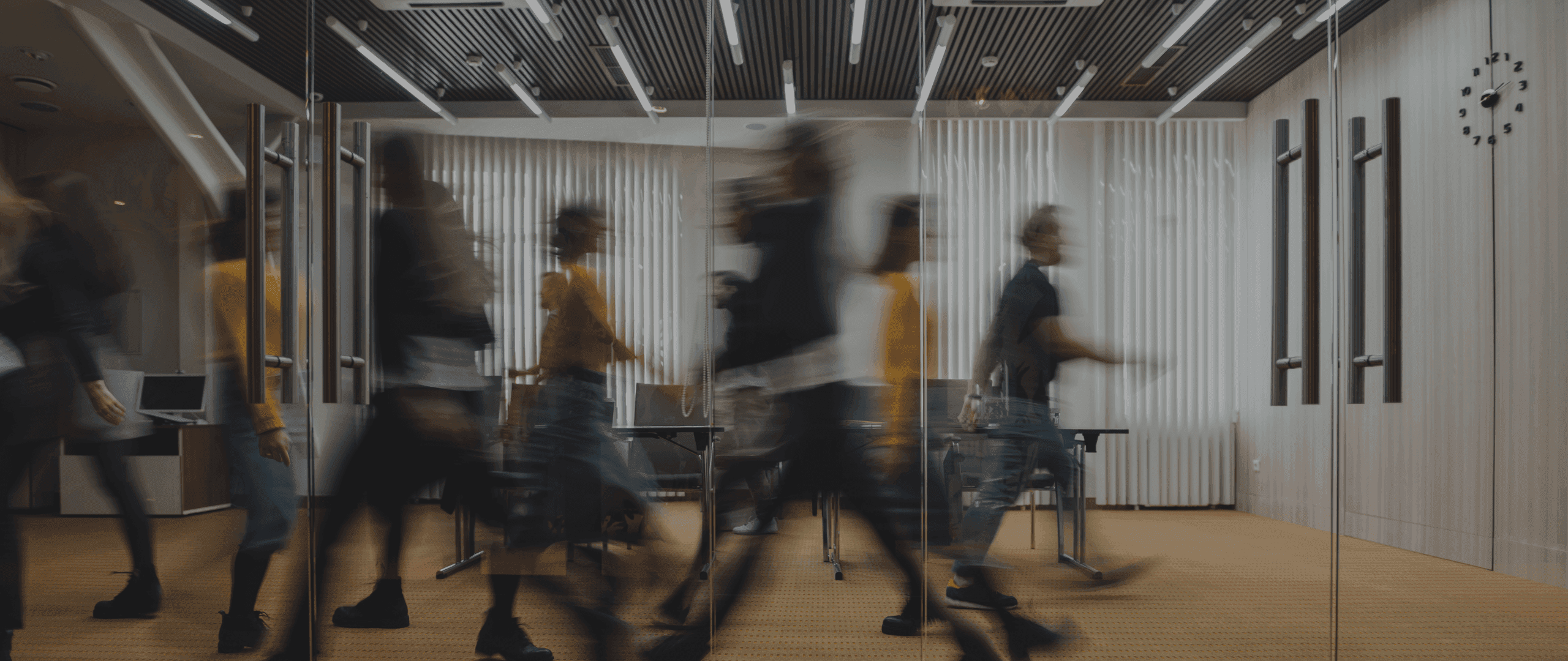 Motion-blurred people walk quickly through a modern office hallway past glass walls.