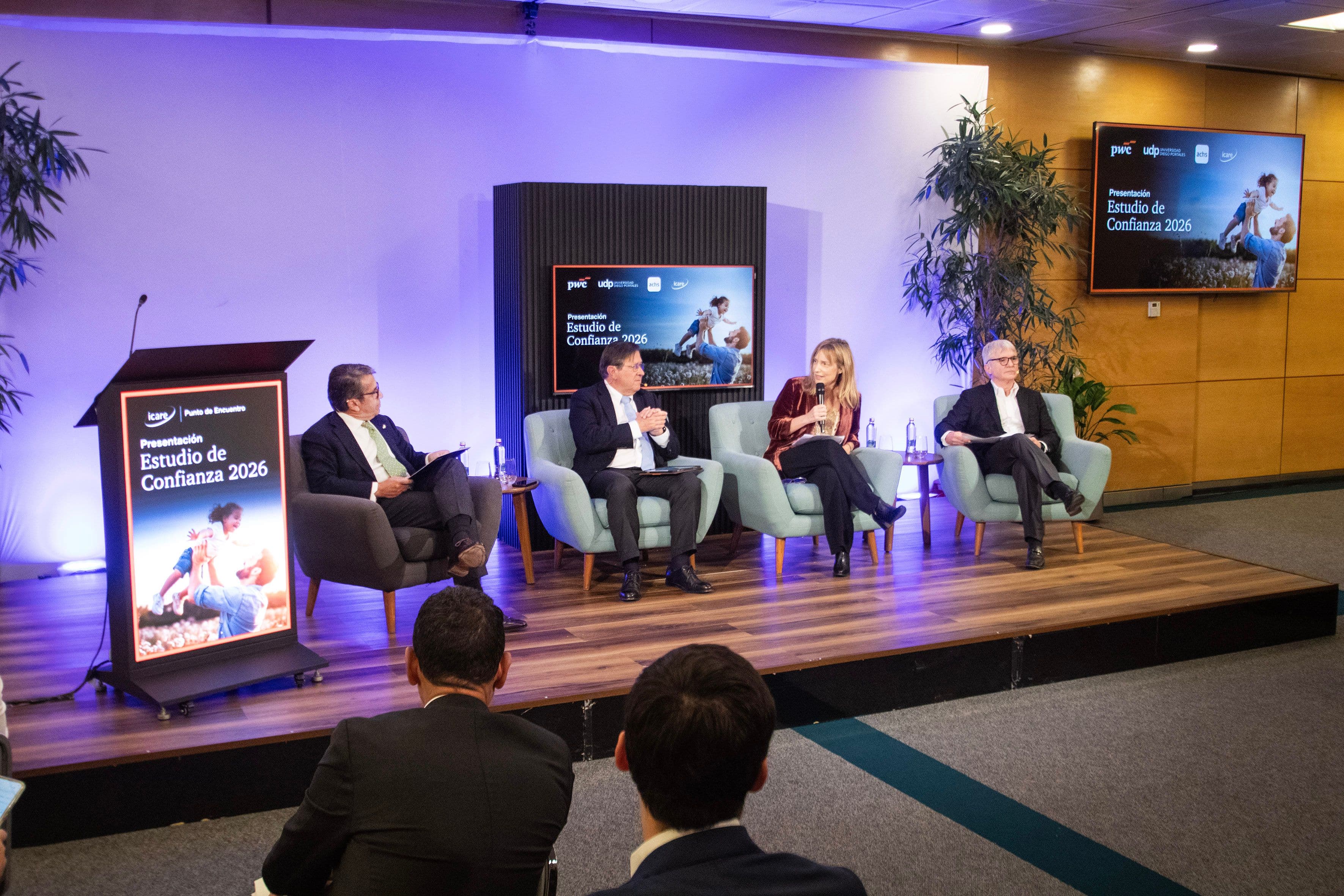 Four panelists engage in a discussion on a stage with screens displaying an event title.