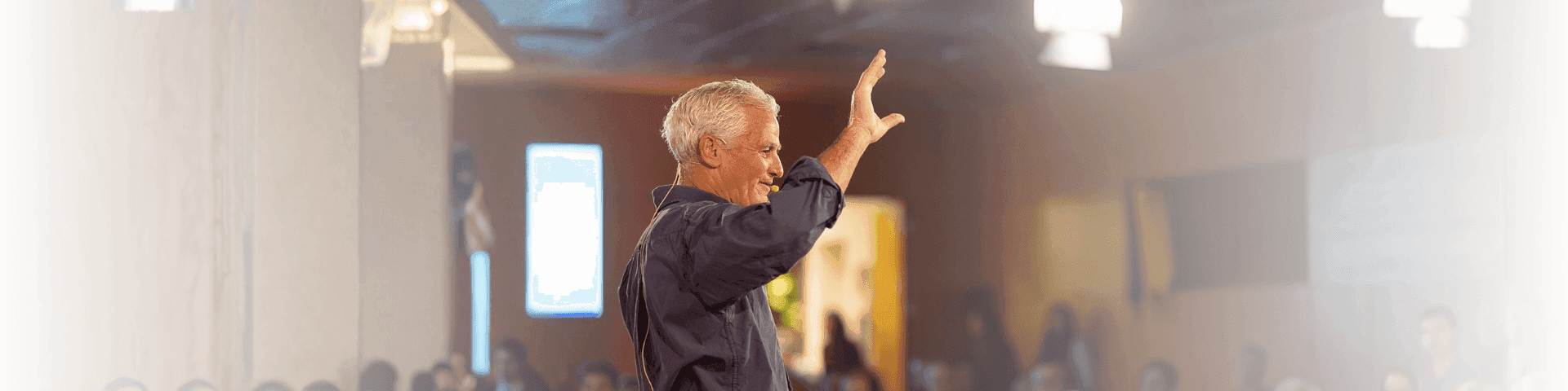 An older man with white hair raises his hand while speaking at a conference.