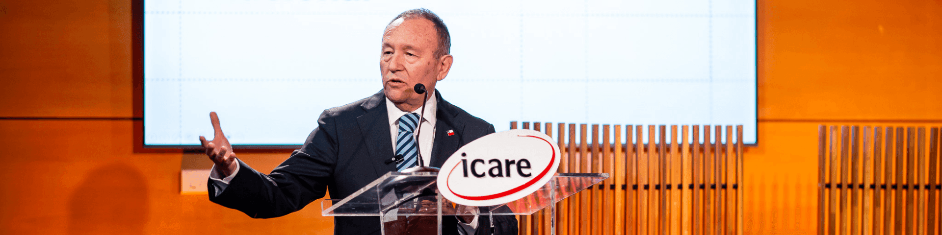Man in a suit speaks at a podium with an 'icare' logo, gesturing with his hand.