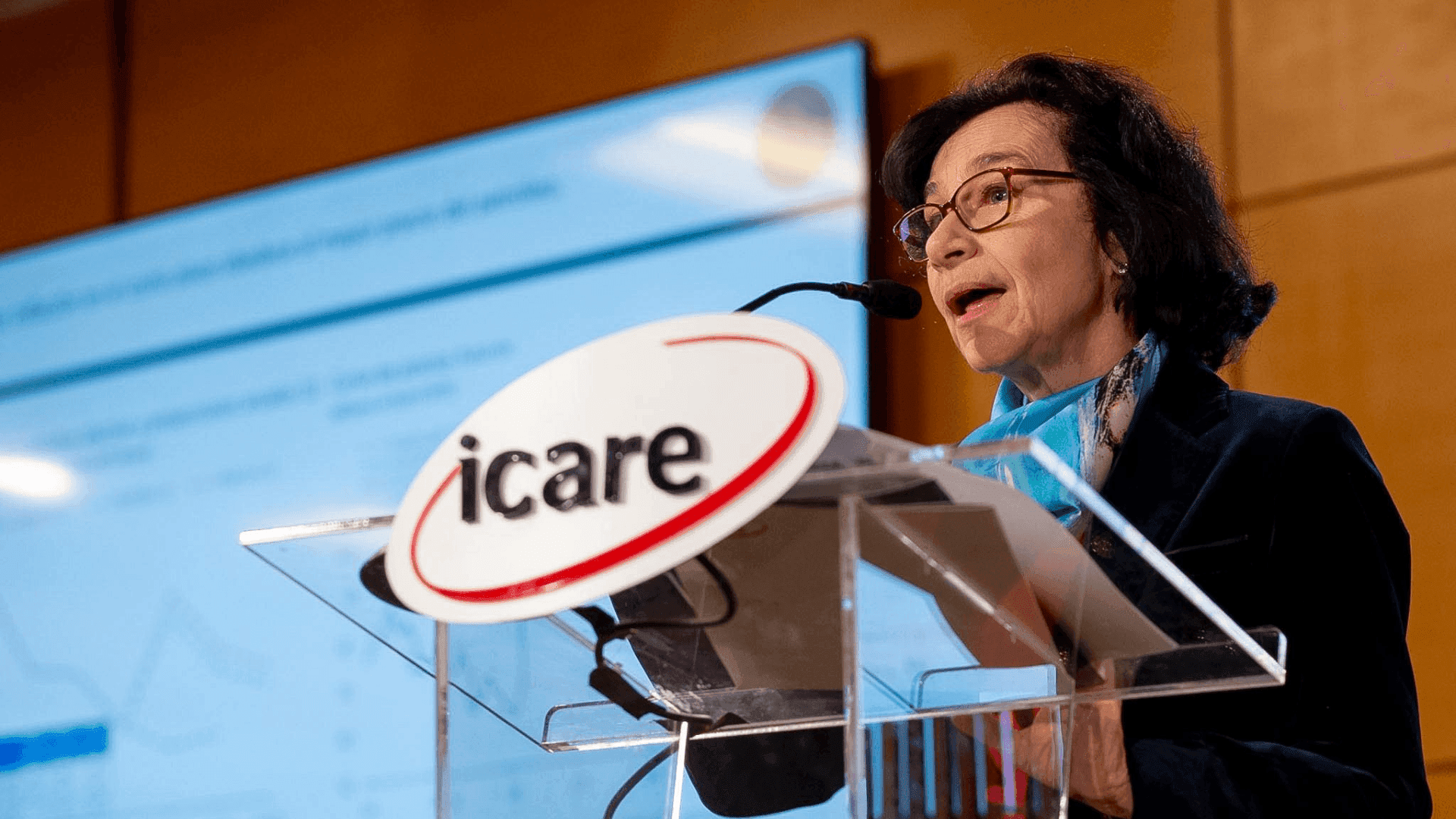 Woman speaks at a clear podium with 'icare' logo, presenting data on a large screen.