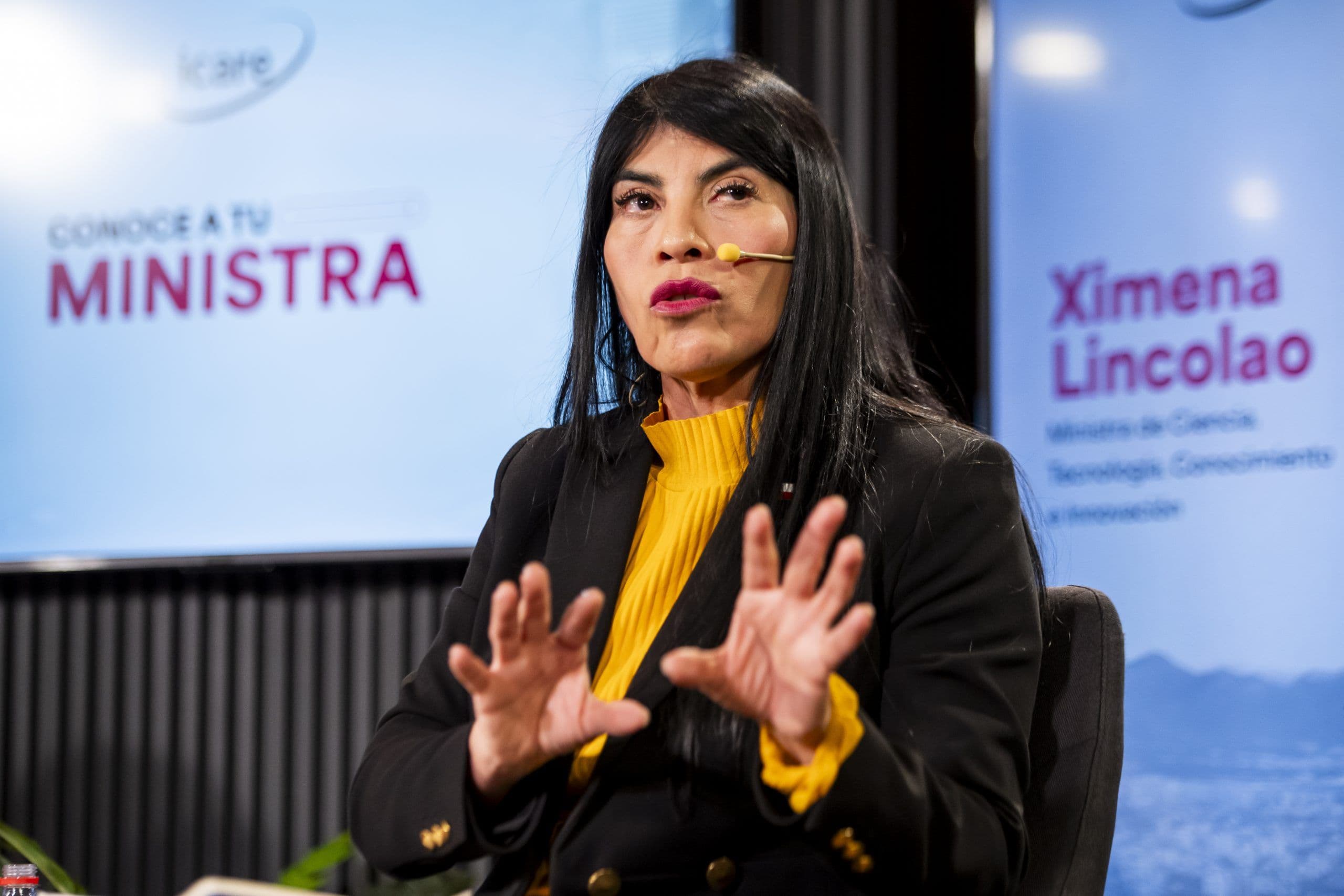 Woman minister gesturing while speaking at a presentation with her name on screen.