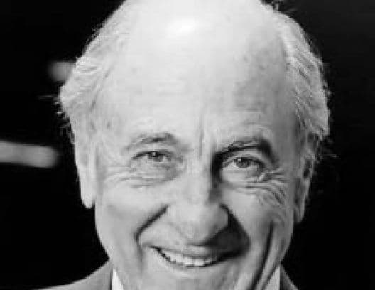 Smiling older man with white hair in black and white portrait, dark background.