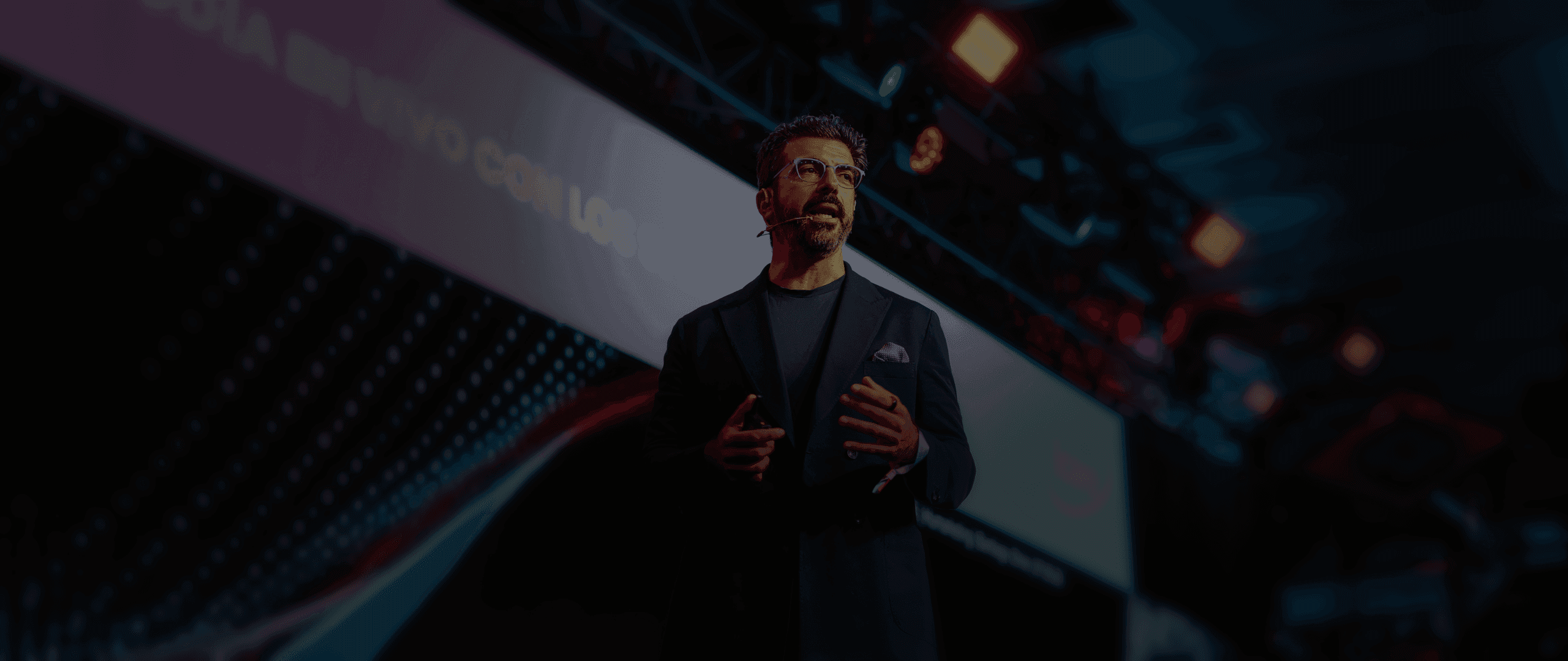 Man speaking on stage with hands gesturing, a large screen behind him.
